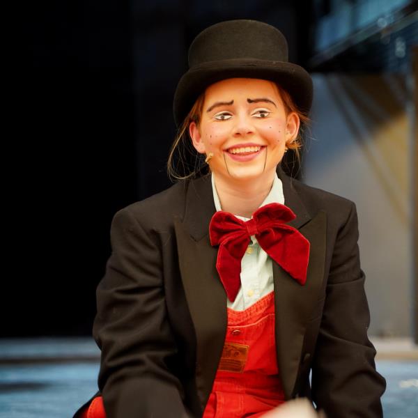 Skuespiller ikledd dress med rød vest og sløyfe, flosshatt på en scene