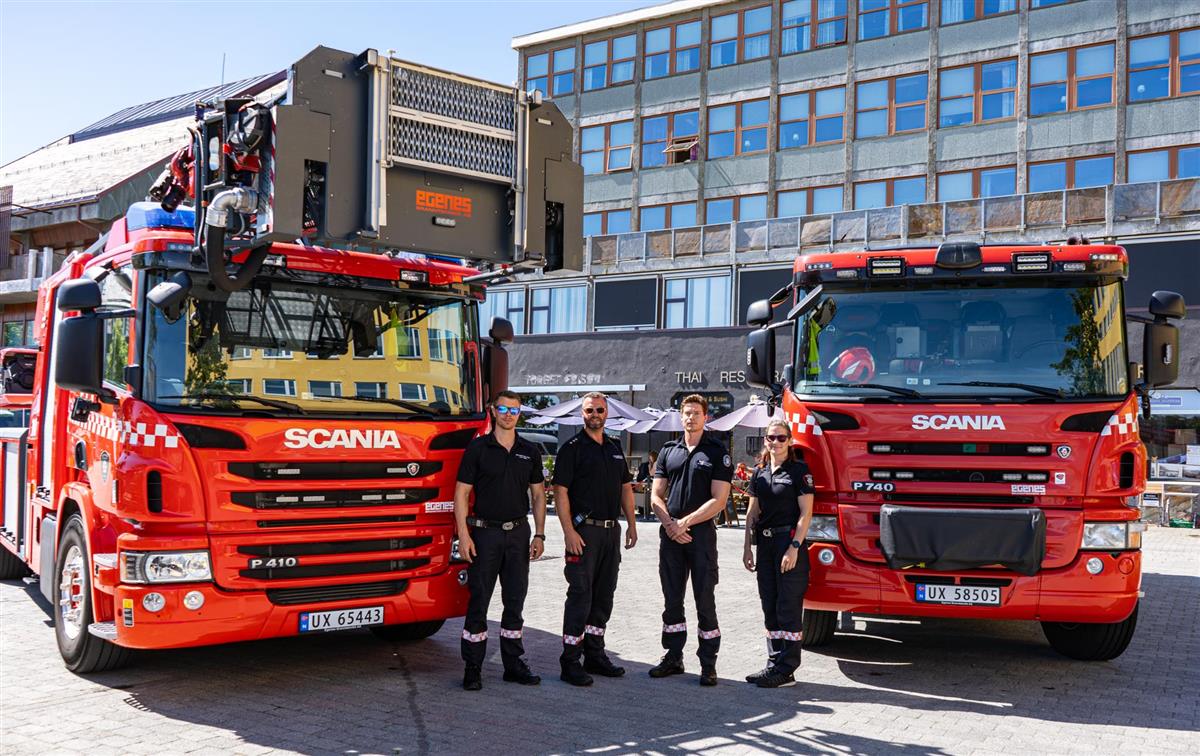 Fire folk fra Nordmøre brann- og redningstjeneste. tre menn og en dame kledd i svart uniform, på rådhusplassen. To brannbiler står rett bak de. - Klikk for stort bilde