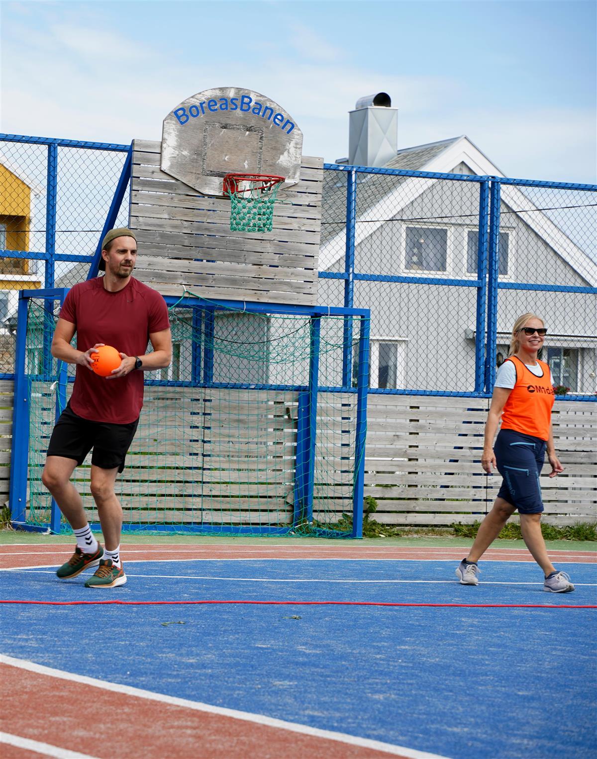 En mann og en dame ikledd shorts, tskjorte og oransje vest kaster ball mot kamera inne i en fotballgrind - Klikk for stort bilde