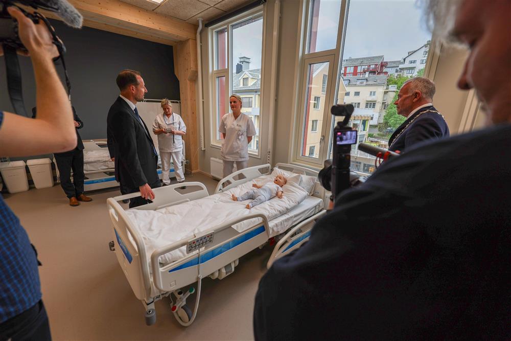 Kronprins Haakon står med sykepleierstudenter og snakker mens de ser på en dukke av en baby i en sykeseng - Klikk for stort bilde